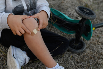 A young boy in a helmet riding a scooter, falls and scrapes his knee. He applies a band-aid to the wound, showing resilience and self-care.