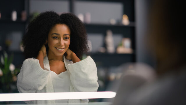 African American happy smiling healthy woman looking at mirror reflection enjoy perfect skin tone after facial mask cream girl pampering touch hair spa procedure at home natural beauty morning routine - Powered by Adobe