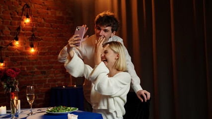 Couple celebrating valentines day and having a romantic dinner in restaurant, sit at table and taking selfie on smartphone, smiling hugging, positive expression.