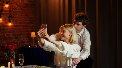 Couple celebrating valentines day and having a romantic dinner in restaurant, sit at table and taking selfie on smartphone, smiling hugging, positive expression.