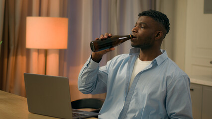 African American man at kitchen male relaxing video call online conference guy alcohol addiction drinking beer bottle home evening bad habit laptop looking at computer watching sport game alcoholism