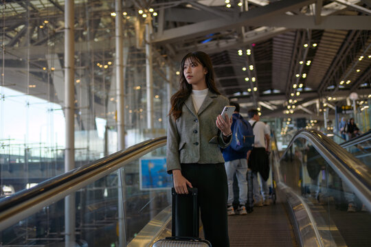 Young businesswoman using her smartphone while ascending an escalator in the airport, managing her trolley suitcase and preparing to board her flight