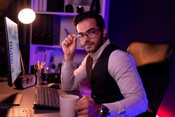 Profile in smart businessman wearing glasses remotely working on desk, looking at camera with waistcoat costume, presenting professional meeting online marketing at neon dark lighting office. Surmise.