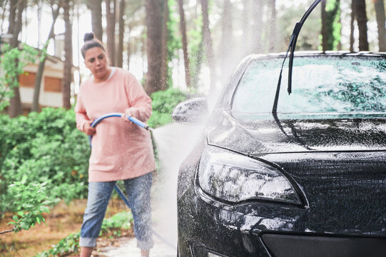 Black Car Is Cleared By Young Woman Using Hose To Spray Water On Driveway Of Her Home