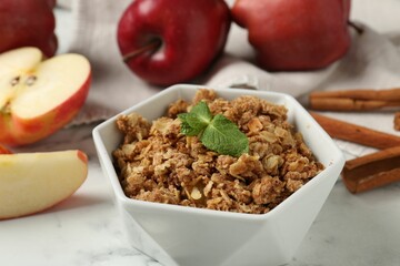 Tasty apple crisp in bowl, fresh fruits and cinnamon on white marble table, closeup