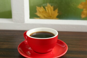 Red cup of aromatic coffee and saucer on wooden sill near window, closeup