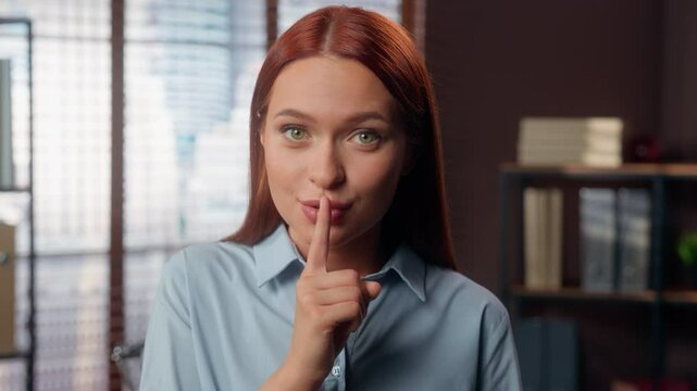 Smiling Caucasian business woman girl female businesswoman employer manager in office hold index finger near lips mouth hush silence quiet gesture secret secrecy information silent looking at camera