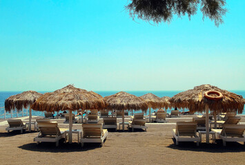 Sun loungers with straw parasols on blue sea background on Perissa beach, Santorini.