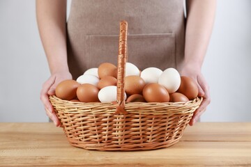 Woman with basket of eggs at wooden table on light background, closeup