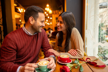 Mid adult Caucasian couple sharing a delightful conversation in a warm cafe environment. Both enjoy lattes at a wooden table, accentuated by a single red rose.