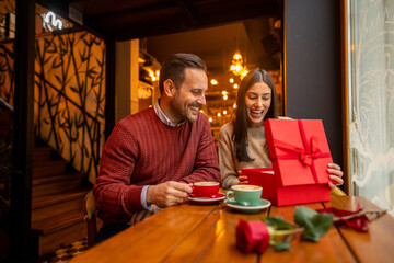 A mid adult Caucasian woman, possibly a professional, joyfully opens a Valentine's Day gift in a cozy cafe, wearing casual attire. The warm setting and a delightful scene perfect for the occasion.
