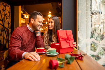 A mid adult Caucasian woman, possibly a professional, joyfully unwraps a Valentine's Day gift in a cozy cafe. She wears casual attire, seated at a wooden table, with coffee cups around her.