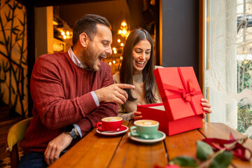 A mid adult Caucasian woman, dressed casually, opens a Valentine’s Day gift with joy. Accompanied...