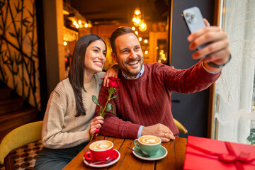 A cheerful Caucasian mid adult couple, dressed in casual sweaters, enjoys taking a selfie in a cozy cafe. The setting features warm lighting, coffee cups, and a romantic gift on Valentine's Day.