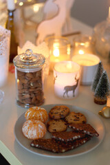 Various Christmas decorations, cookies, chocolate, nuts and tangerines, wine glasses and lit candles on the table. Holiday hygge at home. Selective focus.