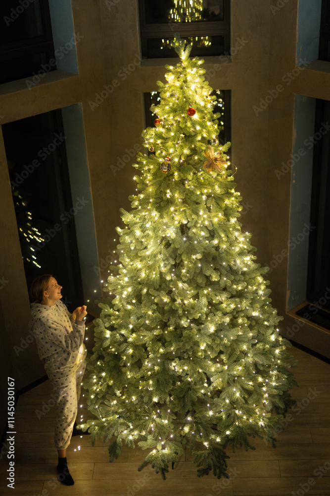 Wall mural A woman in cozy pajamas admires a beautifully decorated and illuminated Christmas tree in a modern indoor setting.