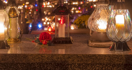 Zapalone świece na nagrobku (i na innych grobach) i róża leżąca na płycie w Dzień Wszystkich Świętych. Dekoracyjne świece i piękna róża zdobiące nagrobek w tle, mnóstwo świec oświetlających cmentarz . © Grzegorz