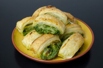Puff pastry rolls with spinach, cream cheese and parmigiana on yellow plate on black table, black background