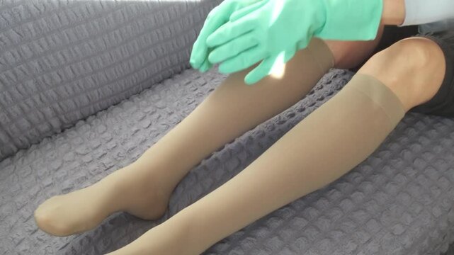An older woman puts on compression stockings at home in the morning. The concept of prevention and treatment of varicose veins, lipedema, spider veins on the legs, leg thrombosis.