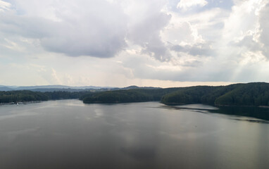 Fototapeta premium Serene lake reflecting the cloudy sky, surrounded by lush forests. A peaceful view ideal for relaxation and nature enthusiasts.