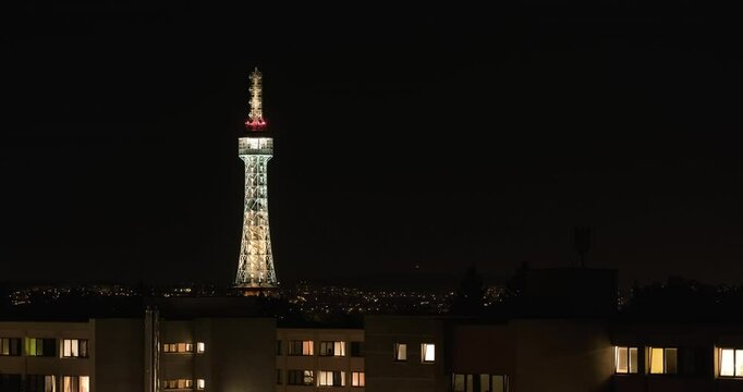 Petř&iacute;n Tower Lit Against Night Prague with Panel Block Windows Flickering Below Distant City Lights and Single Illuminated Plane Performing a Turn Across the Dark Sky Like UFO in 4K Time Lapse