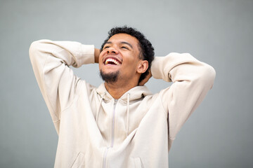 Man in beige hoodie smiling and looking up with hands behind head