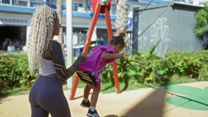 Woman pushing child on swing in city park with urban backdrop and greenery, highlighting playful family interaction and outdoor leisure on a sunny day.
