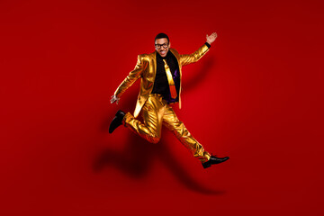 Energetic young man in shiny golden suit jumping with excitement against vibrant red background