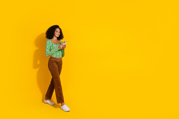 Young woman with curly hair using smartphone in stylish green blouse on vibrant yellow background