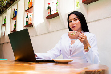 Portrait of a young Colombian woman with cup of coffee in her hands looking at the camera while working on a laptop. Business woman concept