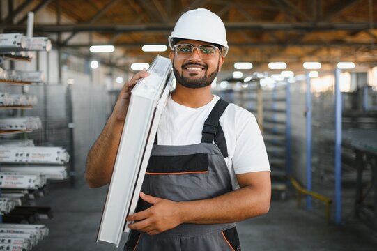 Happy bearded Indian male factory worker in special uniform and white hard hat holding aluminum frame at production of metal plastic windows and doors