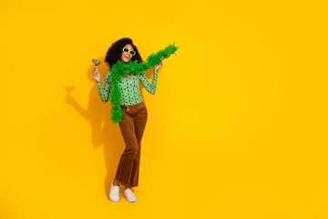 Young woman with black curly hair in vibrant fashion clothes holding a cocktail on a yellow background, exuding joy and charm