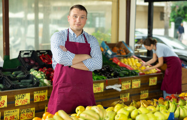 Friendly man seller stands near garden stuff display case and gestures to invite customers to come shopping. Male store employee interrupted work and stands resting and smiling at passing visitors