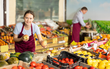 Portrait of smiling female vendor standing near fruits and vegetables stalls at farmers market, offering ripe sweet apples