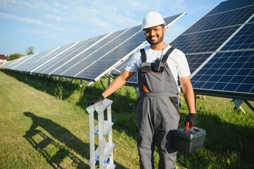 Portrait of Young indian man technician wearing white hard hat near solar panels against blue sky....