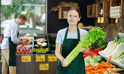 Woman seller of fruit and vegetable store works near showcase with garden-stuff, sorting greenery celery, checking goods.