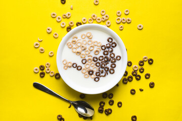 A mix of chocolate and plain cheerios floating in milk in a plain white bowl and scattered on a yellow background. A spoon sits beside the bowl.