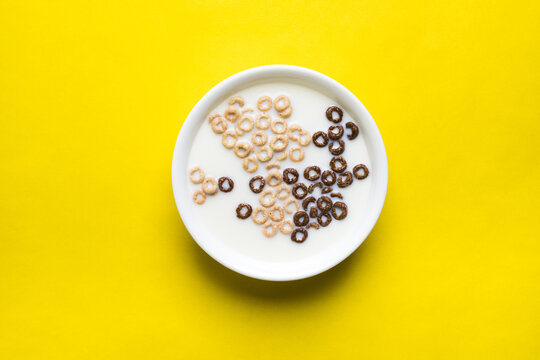 A mix of chocolate and plain cheerios floating in milk in a plain white bowl on a yellow background