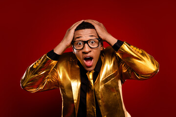 Young man in a golden suit shocked with hands on head against a vibrant red background