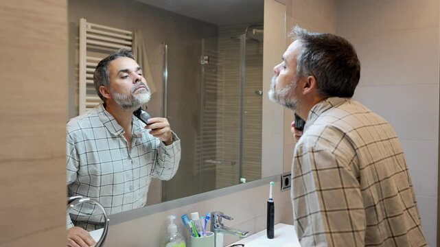 Slow motion sequence of a mature man shaving his beard with an electric razor in front of a bathroom mirror