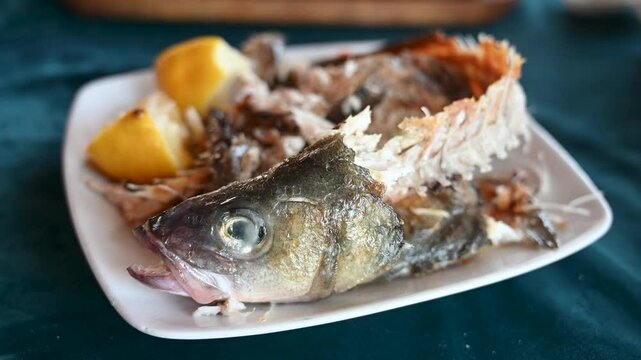 Leftover of the fish. Head and bones of the fish on plate. Dinner leftovers on a table. Remains of Sea bass.