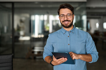 Portrait of smiling male company worker employee, project manager or entrepreneur in office. Happy young professional Latin business man looking at camera standing at work using tablet.