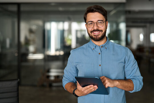 Portrait of smiling male company worker employee, project manager or entrepreneur in office. Happy young professional Latin business man looking at camera standing at work using tablet.