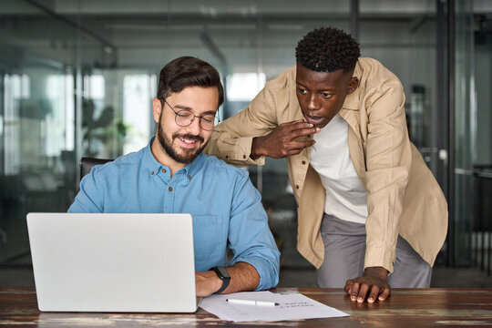 Two busy diverse professional male coworkers software engineers team discussing project together working in teamwork talking sharing ideas sitting at work desk with laptop computer.