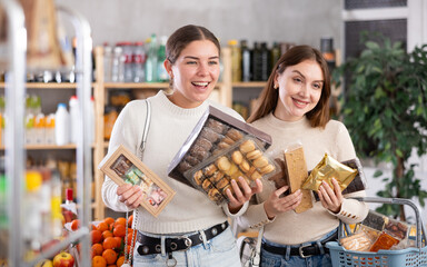 Two European women discussing buying sweets and desserts in a supermarket. Friends choose cookies, chocolate, nougat, for tea. Delicious European sweets