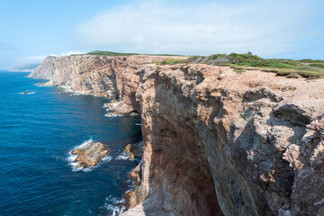 Near a deep blue ocean, a rugged coastline and cliff are in the foreground. It's a bright sunny day with a blue sky and a few clouds. An island with greenery, buildings, and rock is in the back. 