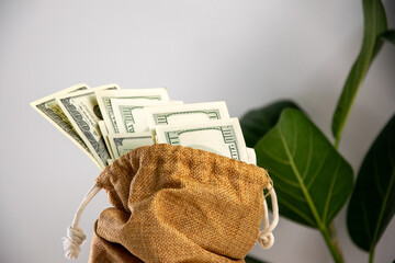 A burlap bag with a drawstring sits on a white surface, filled with US dollar bills that peek out...