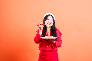 cheerful asian woman biting noodles look left, hand holding fork filled with noodles and white plate, wearing red christmas blouse, Santa hat, isolated on orange background