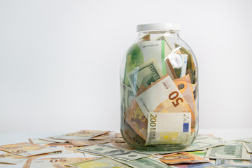 A clear glass jar displays a mix of euro and dollar bills on a white surface. Jars transparency shows currency inside