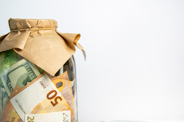 A glass jar filled with dollar and euro bills is covered with brown paper and a rubber band. The jar is on a white background